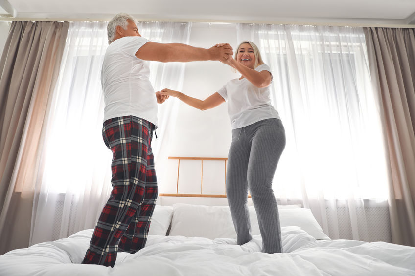Happy mature couple dancing together on bed at home, low angle view
